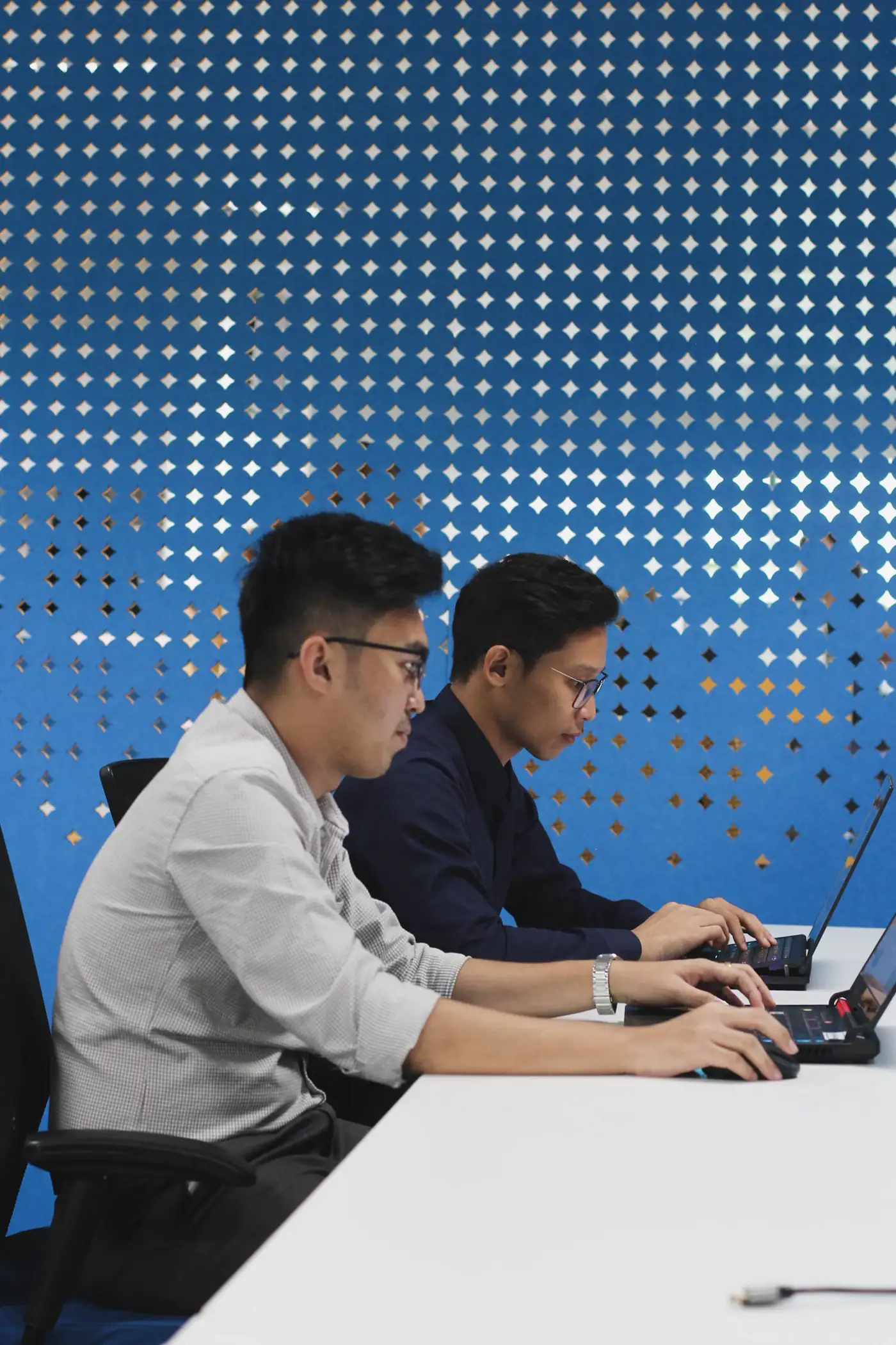 Two people working on laptops in front of a blue perforated wall