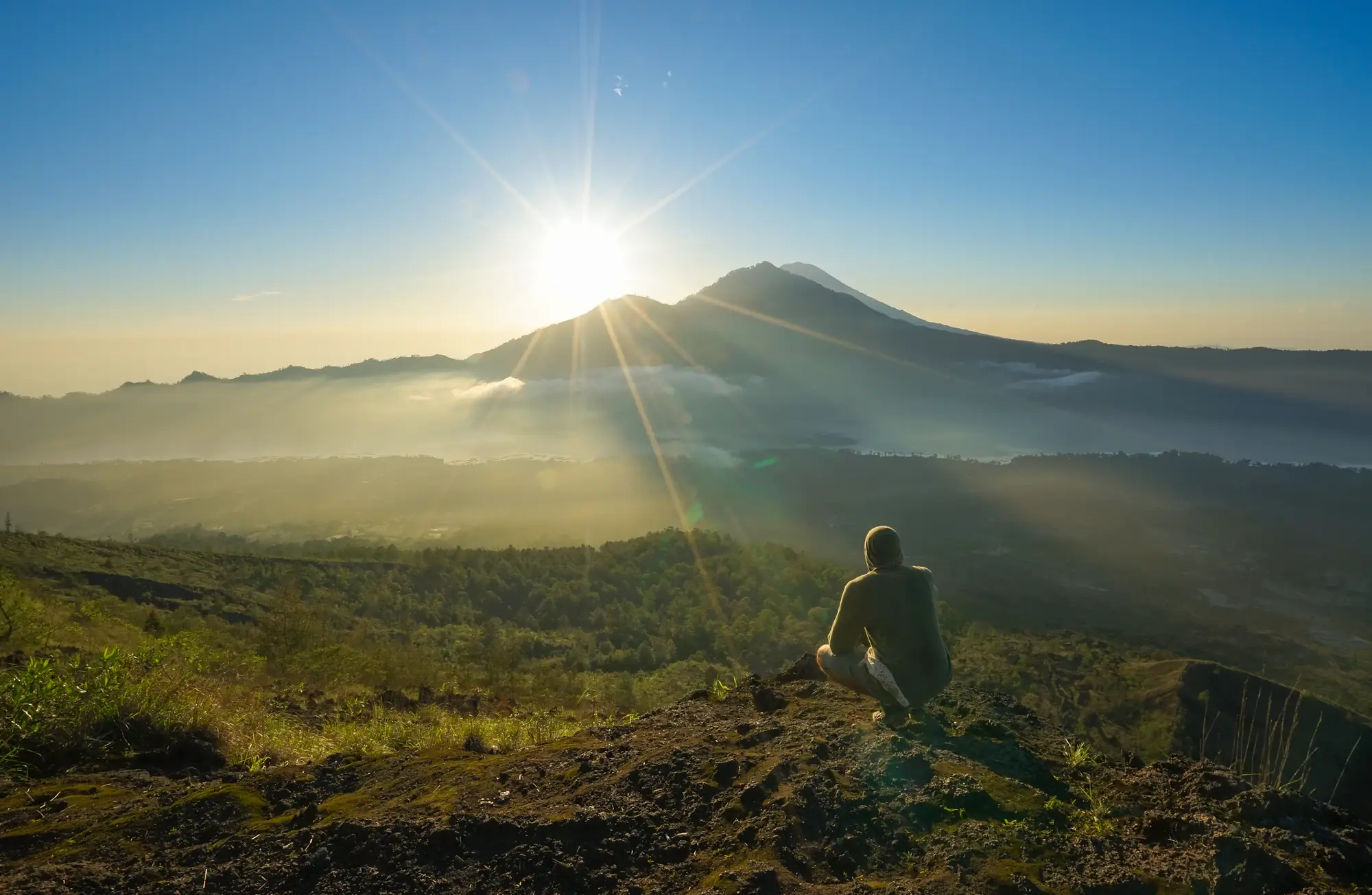 A hiker sitting above the clouds watching the sun rise behind a volcano